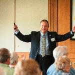 A man holds both arms up while holding a wireless mouse, while two people seated in front of him copy his gesture
